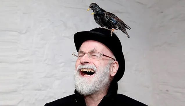 Sir Terry Pratchett is a smiling white man with a white beard, and is wearing a black hat. A black bird perches atop the hat.