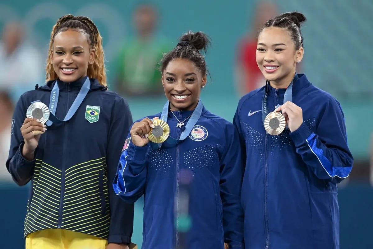 Rebeca Andrade, Simone Biles, and Suni Lee pose at a podium with their Olympic silver, gold, and bronze medals respectively.