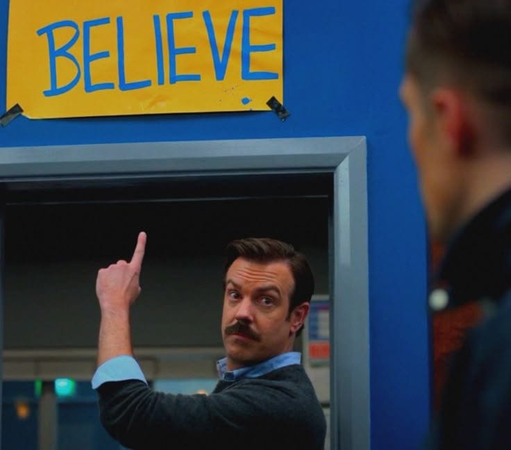 Jason Sudeikis, as Ted Lasso, is a white man with a mustache. He is in front of a doorway and points to a yellow sign that has "BELIEVE" written on it in blue.